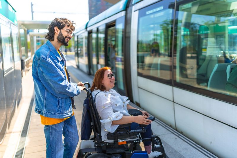 Young man assisting a joyful woman in a wheelchair as she boards a modern tram, highlighting accessibility and support in urban life