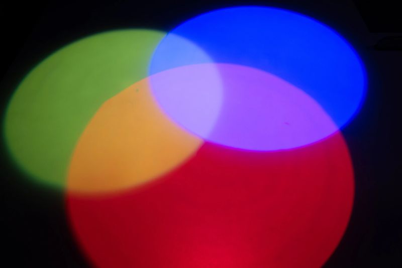 A vivid abstract composition showing the overlap of red, green, and blue light projected onto a dark surface. The intersections create secondary colorscyan, magenta, and yellowdemonstrating the additive color model in a visually striking way.