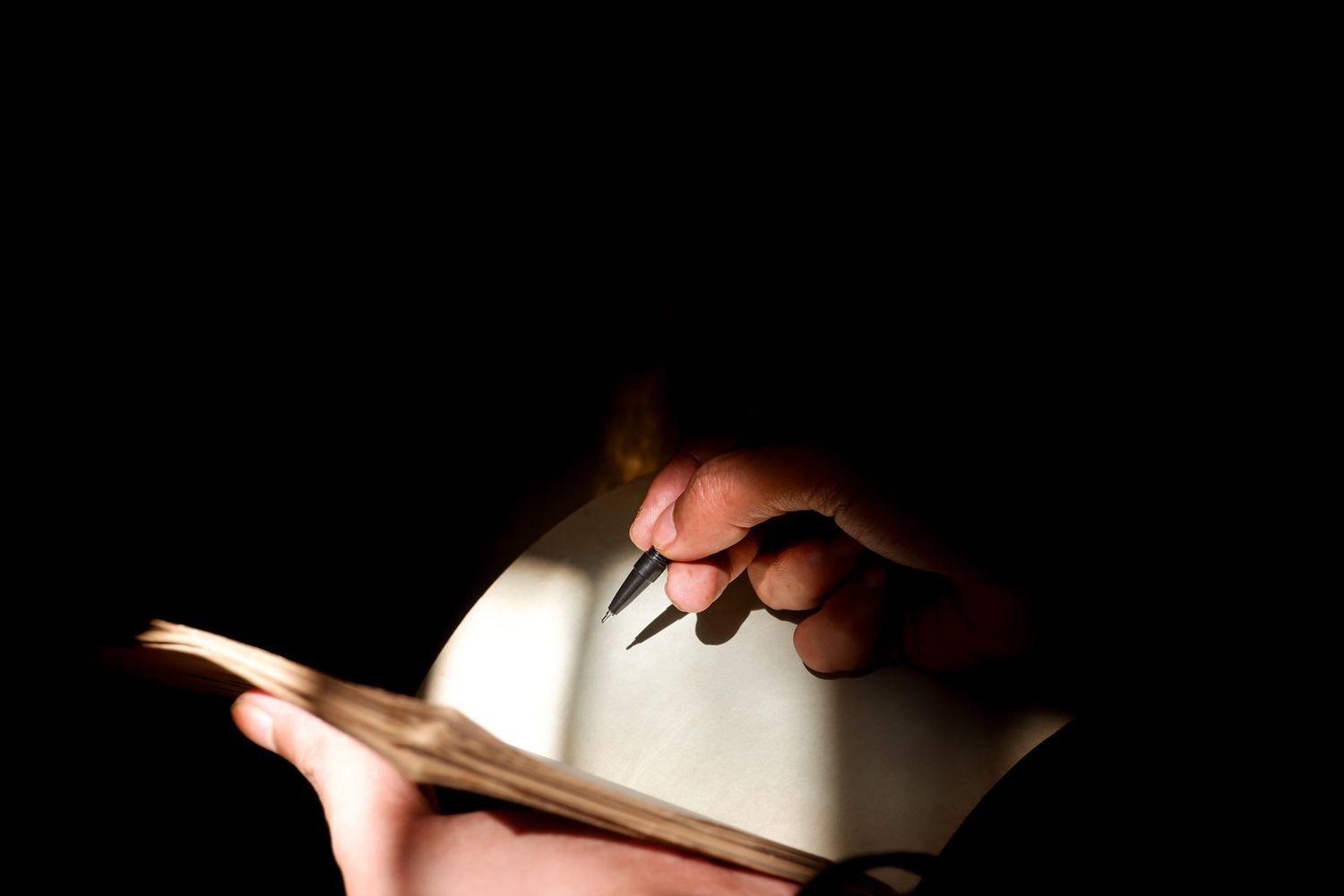 Hand holding a pen poised to write on blank paper in low light.