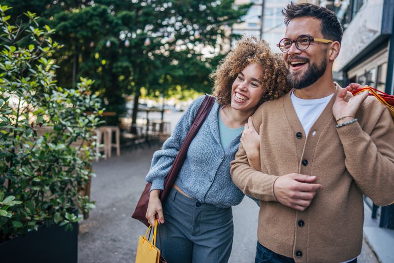 Young couple walking hand in hand down a bustling street, carrying colorful shopping bags and radiating joy after enjoying a delightful shopping spree together