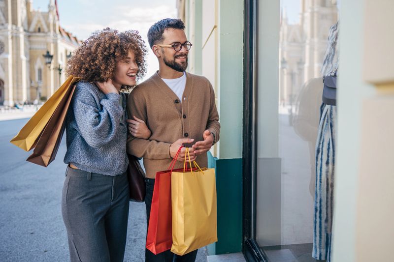 Happy young couple enjoying a day of window shopping, carrying colorful bags while discussing the latest fashion trends they see displayed in the shop window