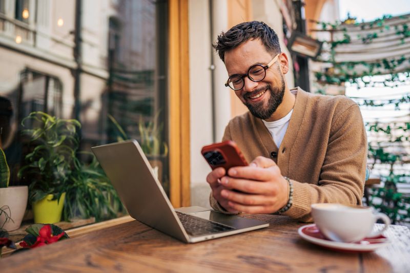 Young adult freelancer smiling while using a smartphone and working remotely on a laptop at an outdoor cafe, enjoying a cozy atmosphere with a pleasant street view