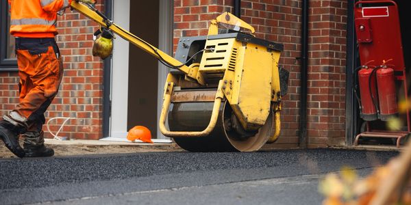 Worker operating road roller on fresh asphalt near building.
