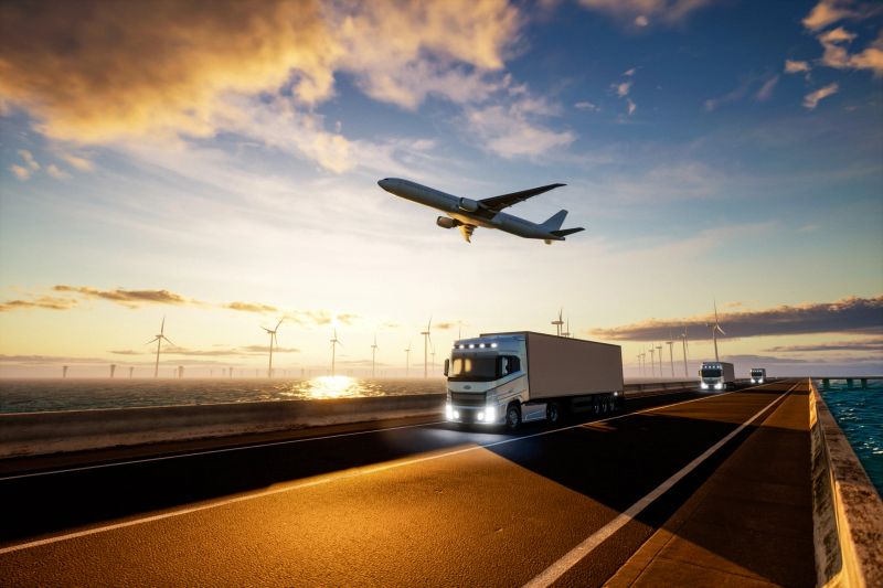 A column of semi-trucks driving along a coastal highway at sunset, with a plane flying overhead and a field of offshore wind turbines in the background. The scene represents the intersection of land, air, and renewable energy transportation, symbolizing a sustainable future in logistics and travel.It is perfect for topics like intermodal green transport, renewable infrastructure, eco-friendly shipping, and clean energy in global logistics.