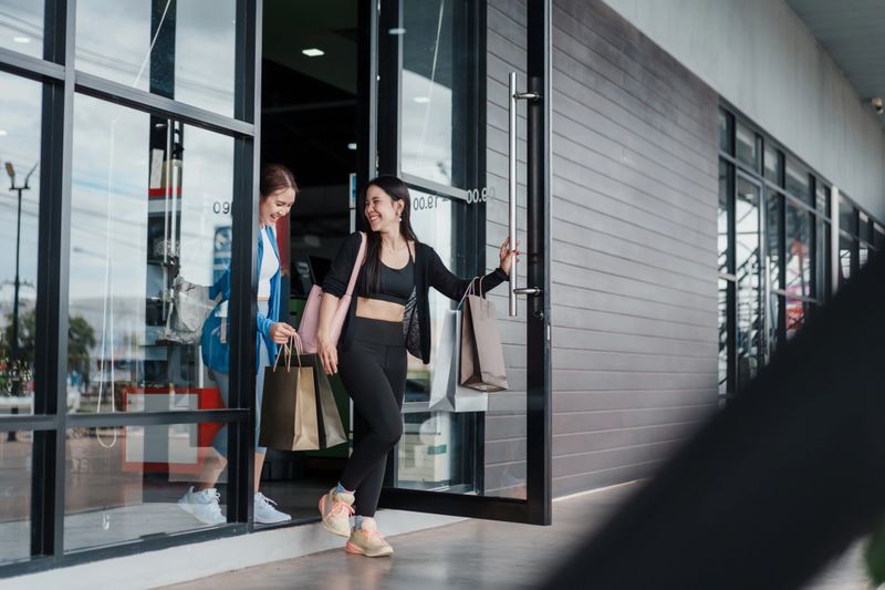 Two happy Asian women exiting sport shop with shopping bags. Friendship shopping moment, fitness fashion, activewear purchase, retail lifestyle, consumer experience, success.