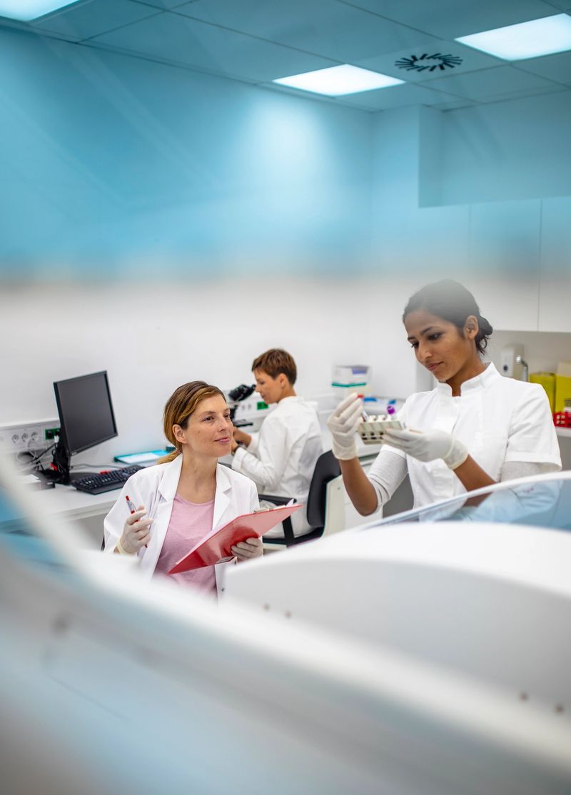 A pregnant Caucasian female scientist collaborates with a young Indian female colleague in a bright, modern laboratory setting. Both are engaged in research tasks, wearing professional lab coats, and using advanced scientific equipment.