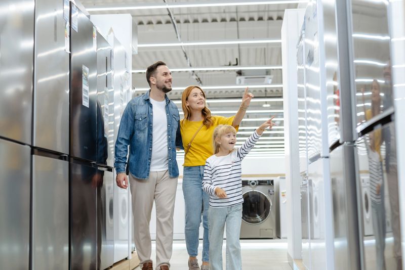 A cheerful family of three explores a retail aisle filled with home appliances, examining products together.
