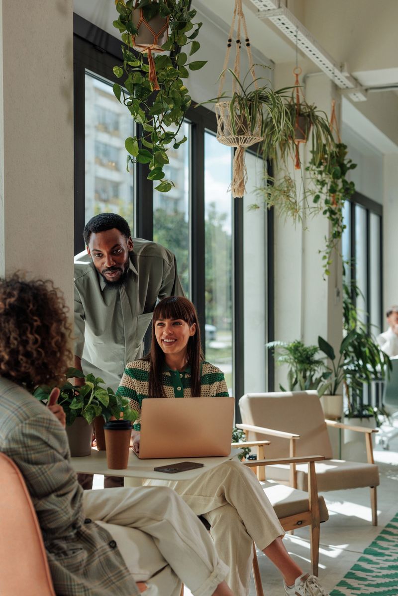 Three colleagues engaging in a lively discussion about a project while collaborating in a modern office filled with vibrant plants and utilizing a laptop