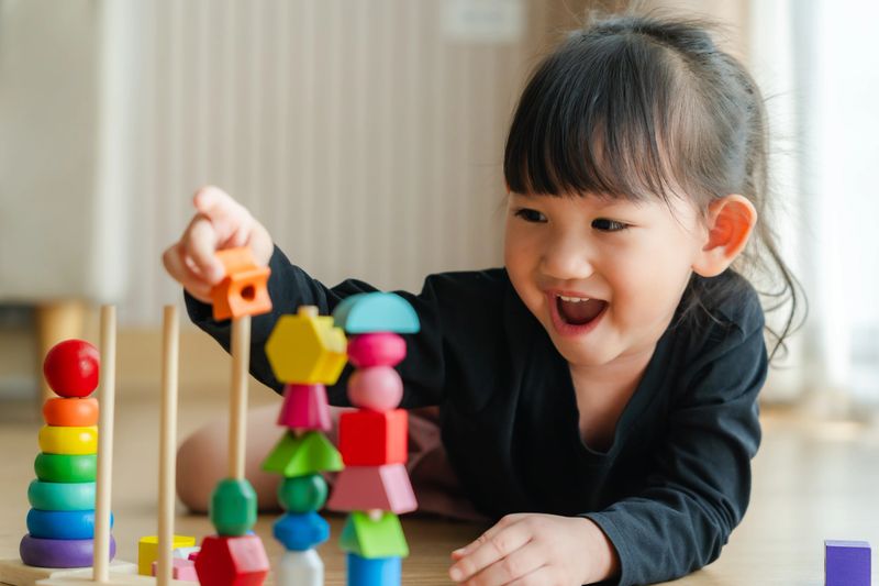 3 year old girl having fun playing with colorful geometric shape wooden blocks educational toys at home. Childhood and kids learning concept.