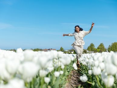 A woman joyfully walks through a field of white tulips under a clear blue sky.