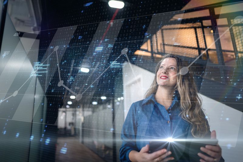 Mature woman wearing a blue shirt and holding a tablet, looking upward with a confident smile. Digital binary code, graphs, and light effects are overlaid to create a futuristic atmosphere, suggesting technology, innovation, and success