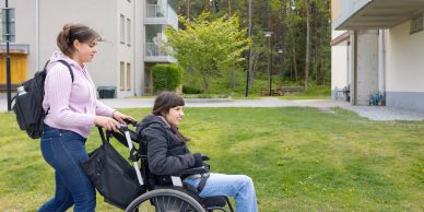Vrouw duwt man in rolstoel buiten nabij appartementen.