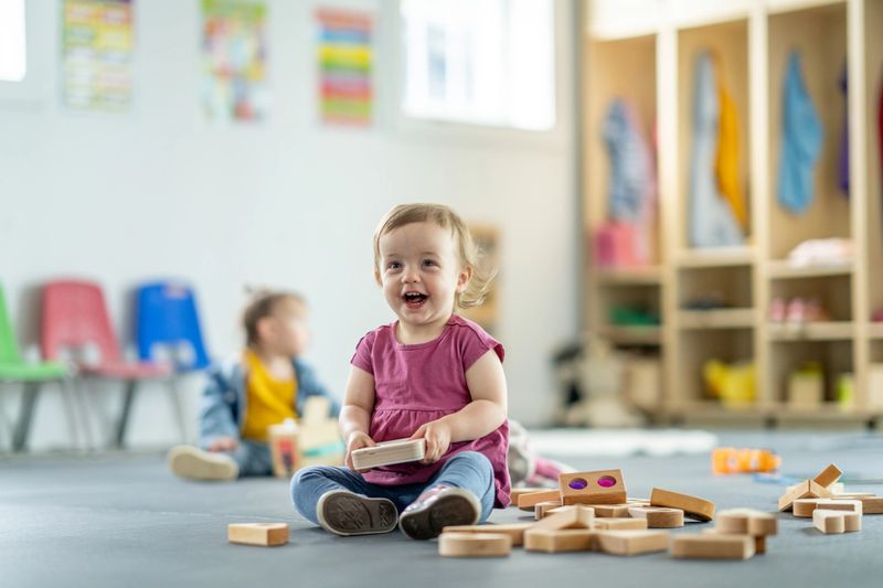 A cheerful toddler sits in a colorful daycare environment, engaged with wooden blocks and sharing joy.