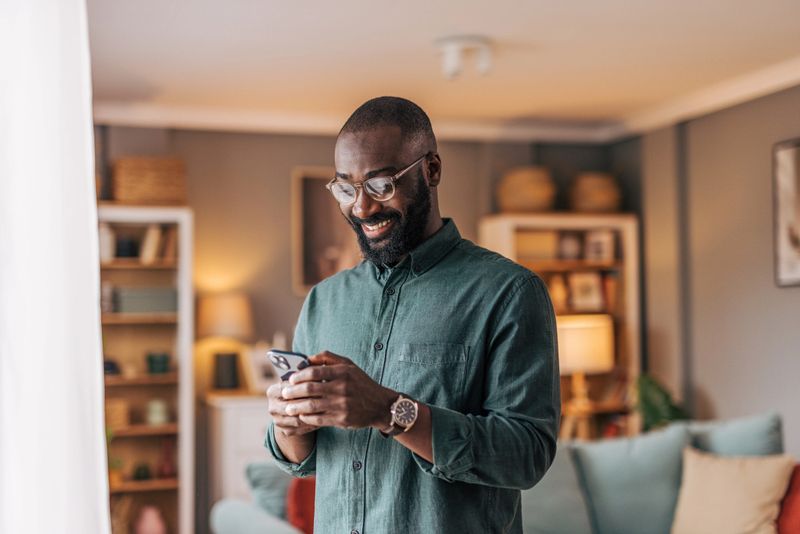 Happy mid-adult man standing in his living room, smiling broadly while reading good news on his smartphone, enjoying a moment of joy and connection in his home