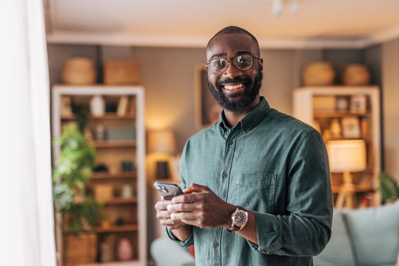 Portrait of a cheerful adult man smiling at the camera while holding a mobile phone in his cozy living room, surrounded by warm decor and natural light, exuding comfort and joy