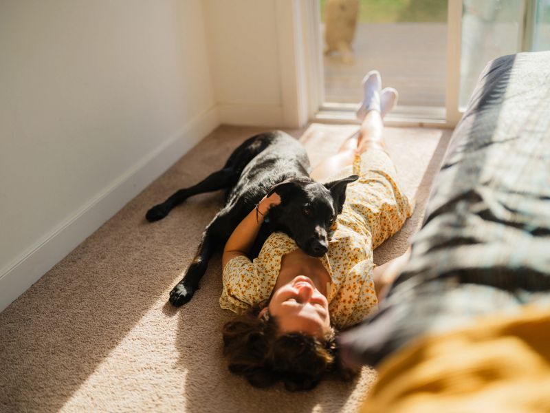 Woman in a summer dress lying on a carpet in a sunlit room near a window, relaxing with her dog. Soft natural light streams through the window, casting warm highlights on the floor. The dog is cuddled up beside her, both appearing calm and content in the peaceful indoor moment.