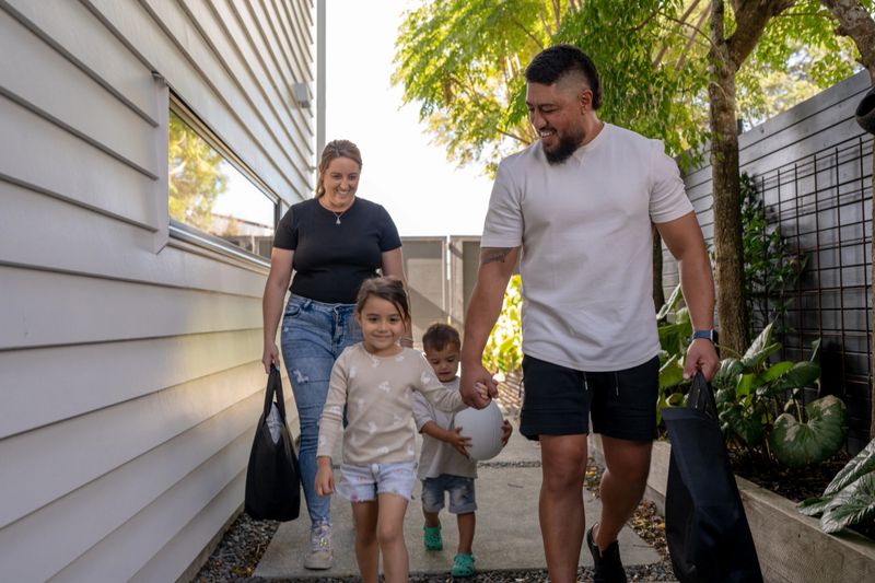 A cheerful family of four walks down a garden path beside their home. The parents carry reusable bags while their children walk ahead