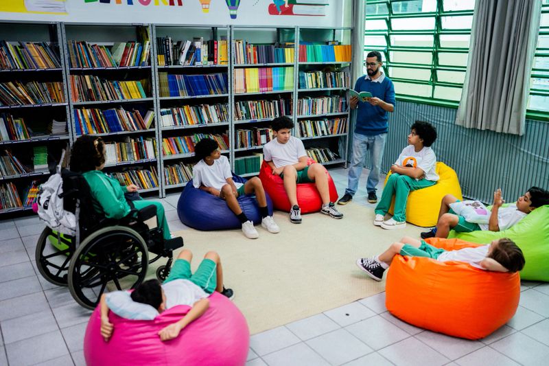 A male teacher stands reading a book aloud to a diverse group of students seated on colorful beanbags in a school library. One of the students is using a wheelchair, reflecting an inclusive and accessible learning environment. The room is filled with bookshelves and bathed in natural light from large windows, creating a warm and engaging space for shared reading and storytelling