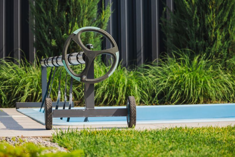 Manual wheeled reel for a pool cover placed beside an outdoor swimming pool with green grass and tall plants in the background. High quality photo