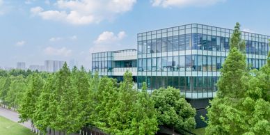 Modern glass office building surrounded by lush green trees under a blue sky.