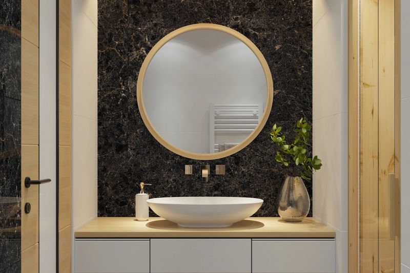 Elegant and compact modern bathroom featuring a one-person pine wood Finish sauna for a cozy, spa-like experience. The space combines luxurious textures with minimalist design, including black marble tiles behind a round mirror, a wall-mounted gold faucet, and an oval counter-top sink placed on a natural pine wood cabinet. Matte white large-format tiles cover the remaining walls and floor, enhanced by warm spotlights and subtle LED lighting. Perfect for promoting compact wellness concepts, modern bathroom design, luxury small-space solutions, interior design, or spa-inspired home features.