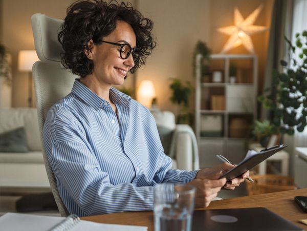 Smiling woman in glasses reviewing documents at her desk in a cozy home office.