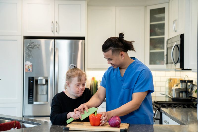 A caregiver in scrubs helps a woman chop vegetables in a modern kitchen. The scene captures moments of support, care, and togetherness, highlighting the value of inclusive collaboration and independence in daily activities.