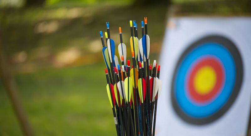 A collection of brightly colored arrows in front of a focused archery target, situated outdoors with a blurred natural background, promoting themes of precision, competition, and recreational sports.