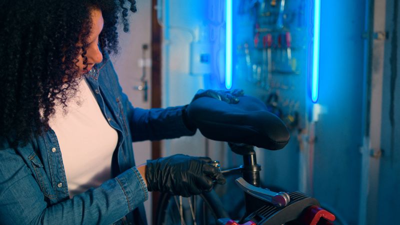 A skilled woman adjusting a bicycle seat in a well-equipped workshop. The atmosphere is enriched with striking blue lighting, highlighting her professionalism and focus on detail during the maintenance process.