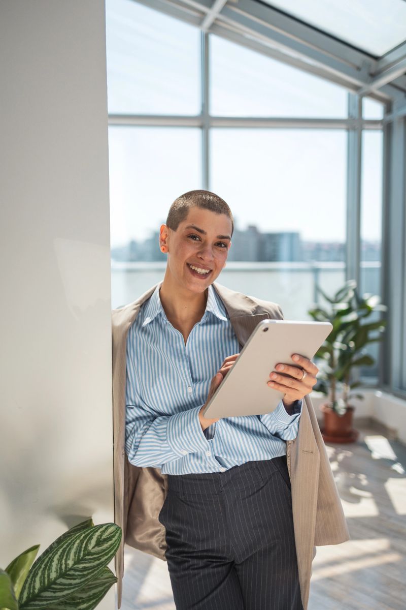 Bright and cheerful professional in a modern office environment, interacting with a tablet device. The setting features natural light, an airy atmosphere, and thriving green plants contributing to the uplifting ambiance.