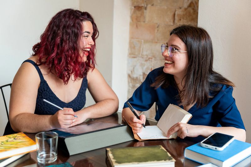 Two young women writers are smiling and collaborating on a writing project, sharing ideas and taking notes during a productive book club meeting
