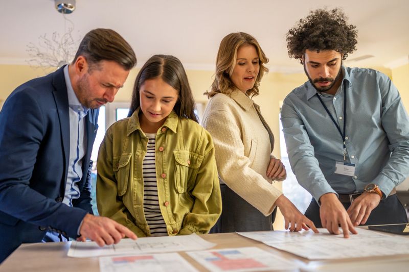 A diverse group including a Caucasian family and Middle Eastern male real estate agent reviewing home floor plans in a brightly lit modern interior.