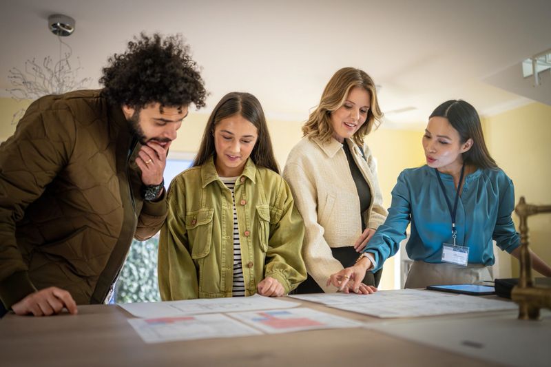 Close-up shot of multicultural family members - mid-aged woman, young adults reviewing real estate documents together at office desk, discussing property details with professional agent.