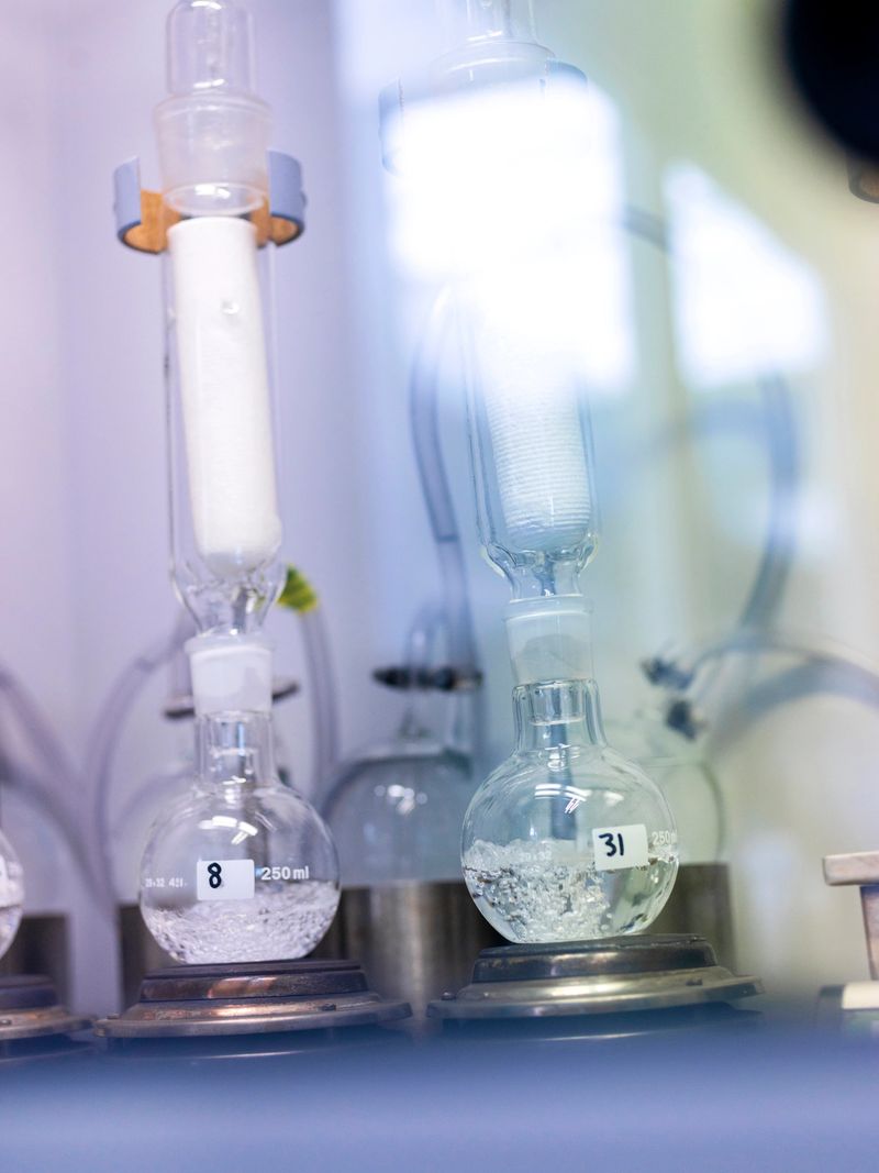 Scientific laboratory setup featuring various glass apparatus including round-bottom flasks, distillation equipment, and numbered sample containers on a laboratory bench. The glassware contains clear liquids and appears to be part of an analytical chemistry or research experiment. Soft lighting and shallow depth of field create an atmospheric view of the professional laboratory environment.
