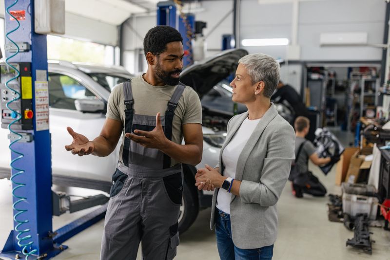 The client is seen seeking clarification from a mechanic in a garage as they discuss underlying car issues, showcasing the importance of effective communication in service.