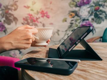 Person holding a teacup near a tablet on a wooden table.