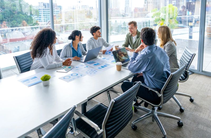 Multi racial diverse group of people working with Paperwork on a board room table at a business presentation or seminar. The documents have financial or marketing figures, graphs and charts on them. One of the businessmen is talking. There are laptops and digital tablets on the table