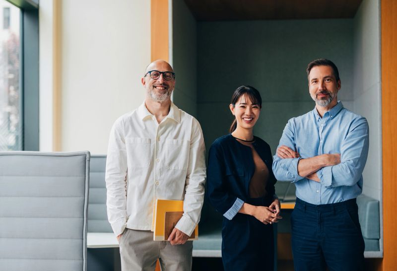 Three professional colleagues smiling and standing confidently in a contemporary office space, symbolizing teamwork, collaboration, and a productive business environment.