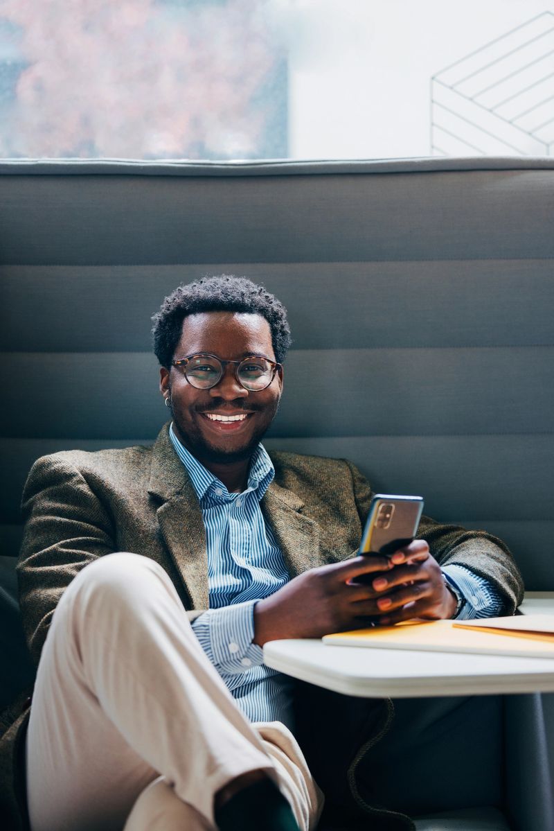 A young business professional smiling while holding a smartphone and seated in a modern office. The ambient environment suggests a workplace setting with a cheerful and confident demeanor.