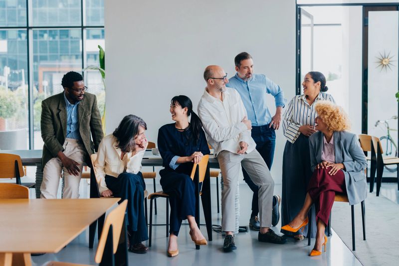 A diverse group of professionals engaging in a cheerful discussion in a well-lit office. The atmosphere exudes teamwork, collaboration, and positivity, reflecting a modern workplace environment and professional camaraderie.