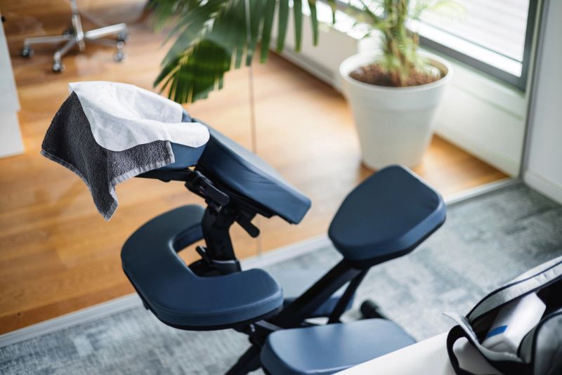 Close-up composition of sleek navy blue ergonomic massage chair with white sanitary cover, positioned on gray carpet near potted palm plant in sunlit office environment.