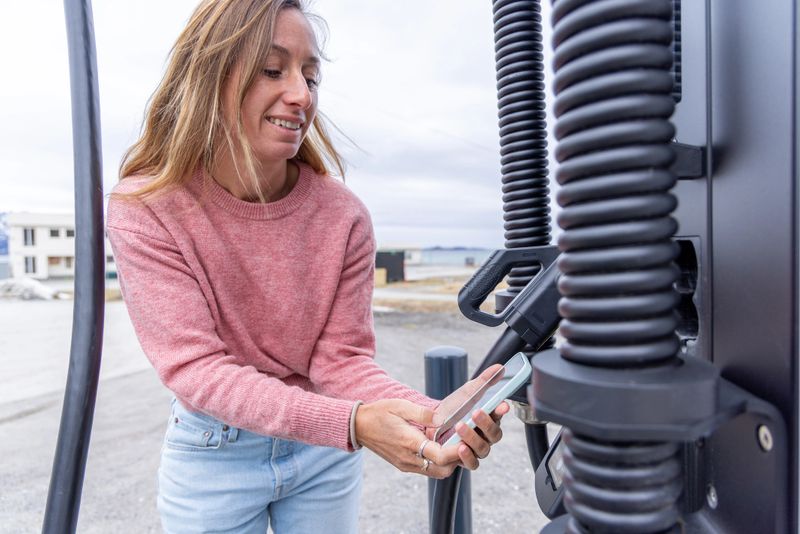 Woman paying with NFC card at electric vehicle charging station. Mobile technology for sustainable transport. Eco-friendly travel with contactless payment.