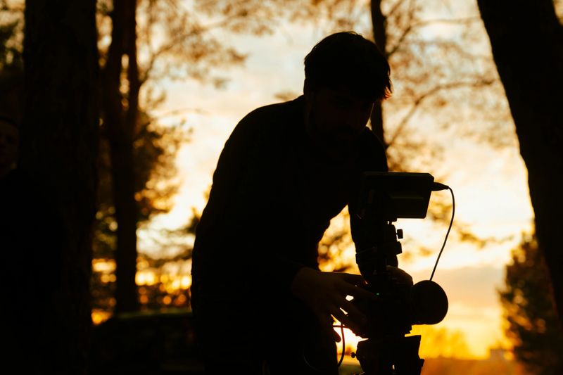 Backstage. A cameraman adjusts equipment in a forest during sunset. The silhouette creates a dramatic scene.