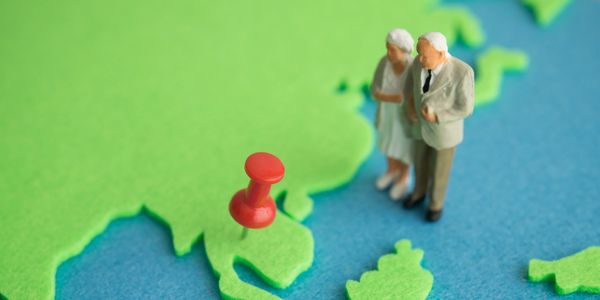 Miniature elderly couple stands near a red pushpin on a map.
