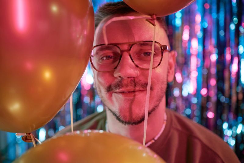 Portrait of young adult Caucasian man with glasses smiling and looking into camera surrounded by balloons celebrating birthday against colorful festive backdrop