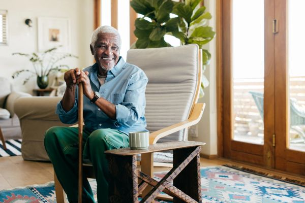 Elderly man sitting with a cane, smiling in a bright living room.