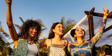 Three women enjoying a sunny day outdoors with arms raised joyfully.