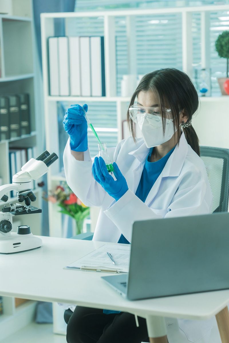 Female doctor in lab coat and gloves pouring blue liquid chemical from plastic bottle into beaker during microscope experiment in science laboratory.