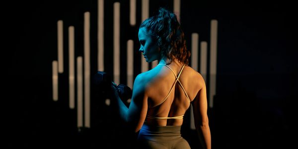 Woman lifting dumbbells in a dimly lit gym with vertical light bars.