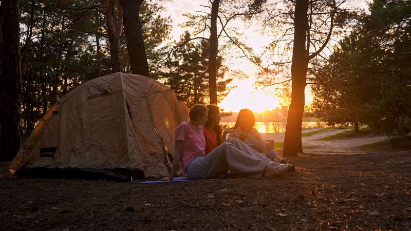 Three friends sit by a tent in the forest during sunset, enjoying the warm glow of the evening sun.